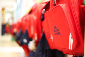 Row of red school backpacks hanging on a rack, with one close-up showing the text 'THE ROCHE SCHOOL' on the front pocket