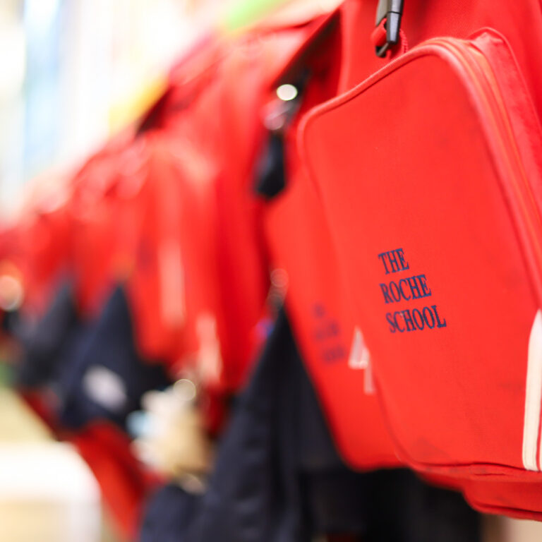 Row of red school backpacks hanging on a rack, with one close-up showing the text 'THE ROCHE SCHOOL' on the front pocket