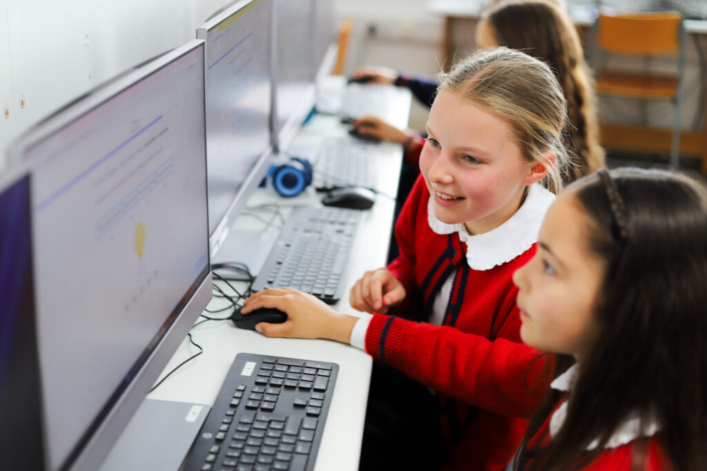Two girls in a computer lab using desktop PCs with keyboards and mice on a long white table.