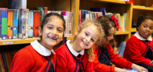 Four schoolchildren in red uniforms sit together on the library floor, reading colorful picture books from an open spread-out book in front of them.