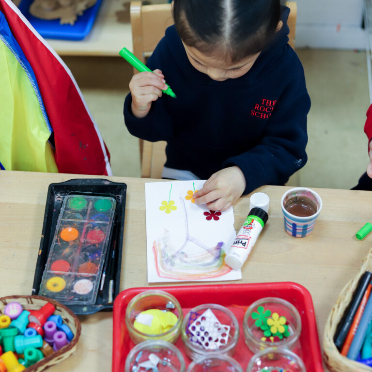 Young child with two red hair ties and a dark hoodie works on a craft project, drawing with a green marker at a table surrounded by paints and supplies.