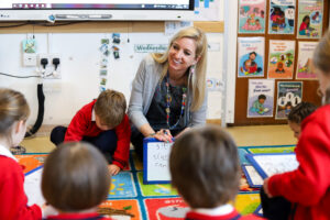 Teacher kneels beside a student at a whiteboard during a classroom activity, smiling as they work together.