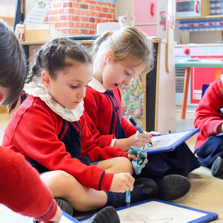 Group of young students in red uniforms sitting on the floor, drawing on blue clipboards in a classroom. (Informative)