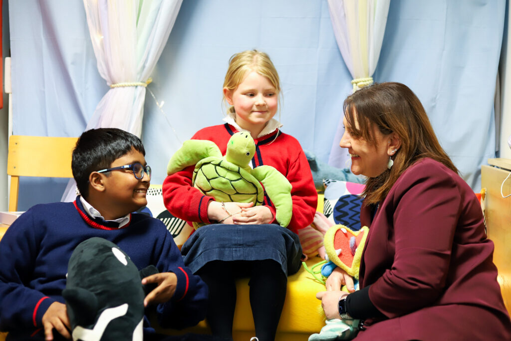 Teacher and two children sit on a yellow couch, smiling at a green turtle plush toy between them in a cozy classroom setting.