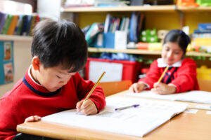 Close-up of a young boy in a red sweater drawing with a pencil at a desk; a classroom scene with a girl at another desk in the background is blurred behind him.