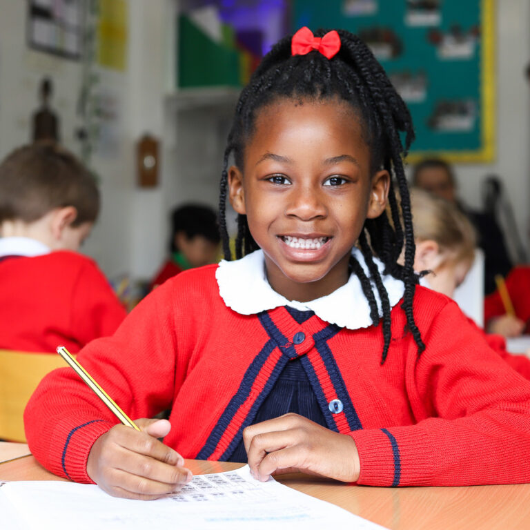Girl in a red sweater smiling at the camera while writing at a desk in a classroom.
