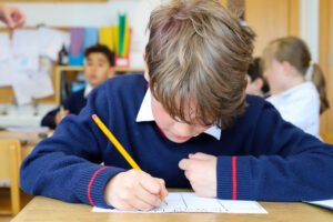 Child wearing a blue sweater writes with a pencil at a desk in a classroom, grid paper in front and colorful materials in the background