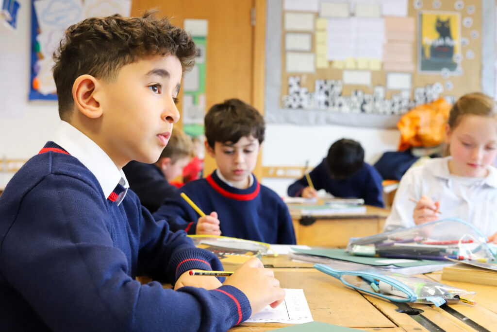 Close-up of a student in a blue sweater writing with a pencil at a classroom desk, other kids in background.