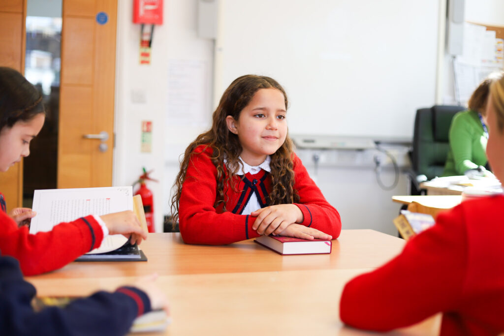 A young girl with long curly hair in a red sweater sits at a table in a classroom, looking thoughtful with a notebook in front of her.
