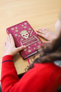 Child hands hold a maroon book titled 'Tales from Shakespeare' with a skull-and-crown design on the cover, on a wooden table.