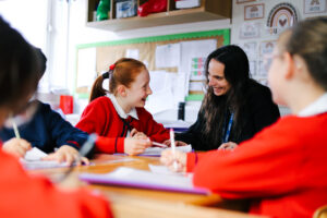 Red-haired student in a red uniform chats with a smiling teacher at a classroom table during a group activity.