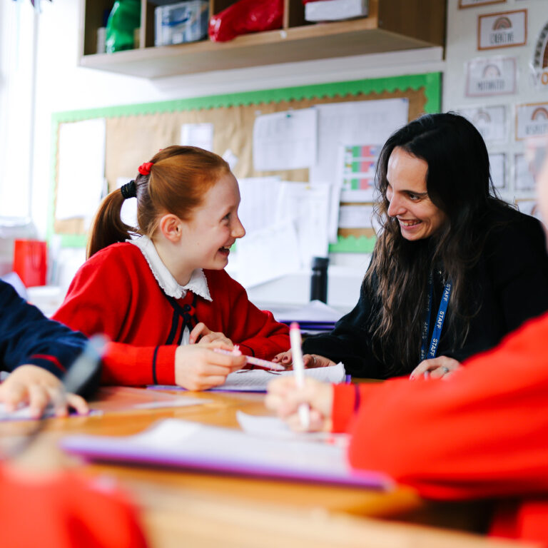 Red-haired student in a red uniform chats with a smiling teacher at a classroom table during a group activity.