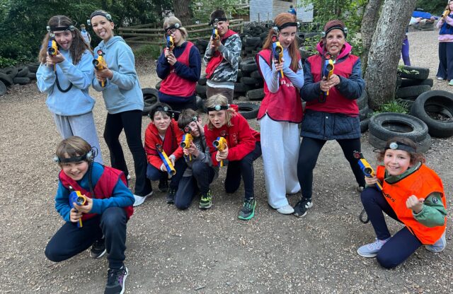 Group of kids with headlamps posing outdoors, holding yellow toy blasters, tires stacked in the background.