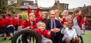 Group of smiling schoolchildren in red sweaters and striped uniforms posing with a man in a suit during outdoor playtime on a playground.